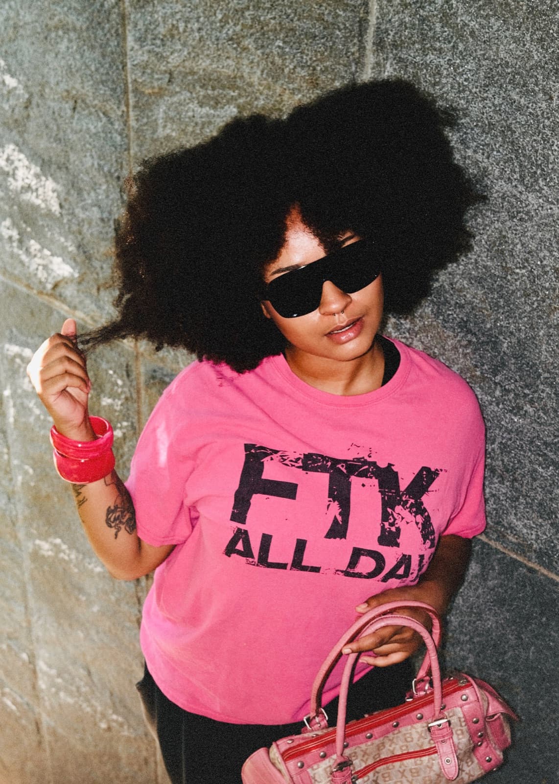 Portrait de Supremnova, lunettes noires et t-shirt rose, contre un mur de pierre